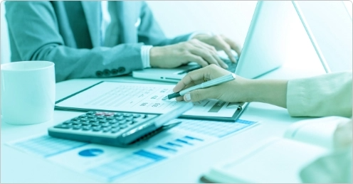 Two people working at a desk with a laptop, papers, and a calculator, suggesting financial or accounting tasks.