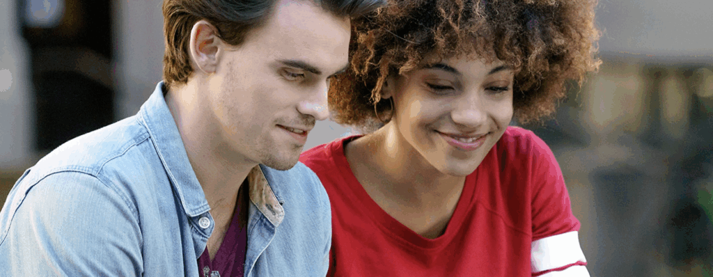 Young couple smiling and talking; man with short dark hair in a denim jacket, woman with curly reddish-brown hair in a red t-shirt.